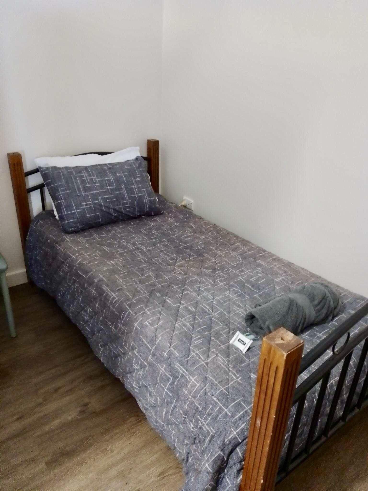 Ein kleines Zimmer mit einem Einzelbett, einer grau gemusterten Bettdecke, einem Holz-Kopfteil und einem grauen Handtuch auf dem Bett.