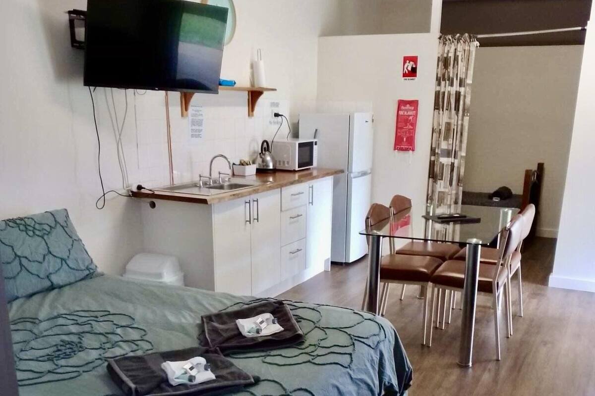Habitación de hotel compacta con cama, kitchenette pequeña, zona de comedor y televisor montado en la pared.