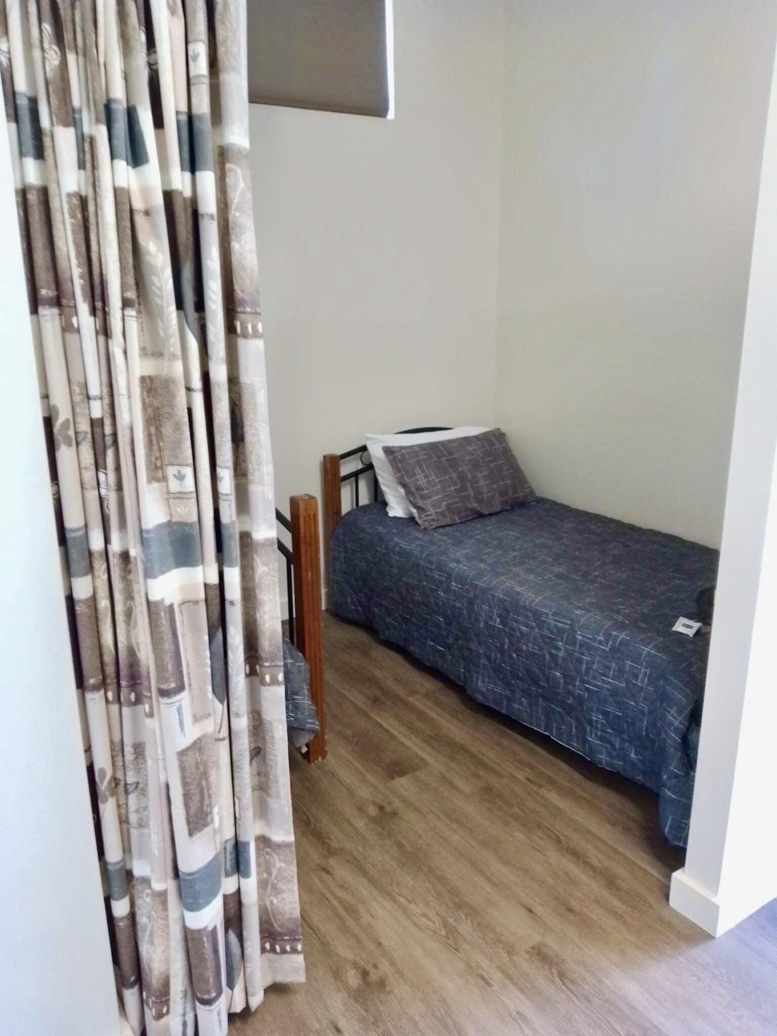 Une petite chambre avec un lit simple, un plancher en bois, un cadre de lit en métal et une couverture de lit grise.