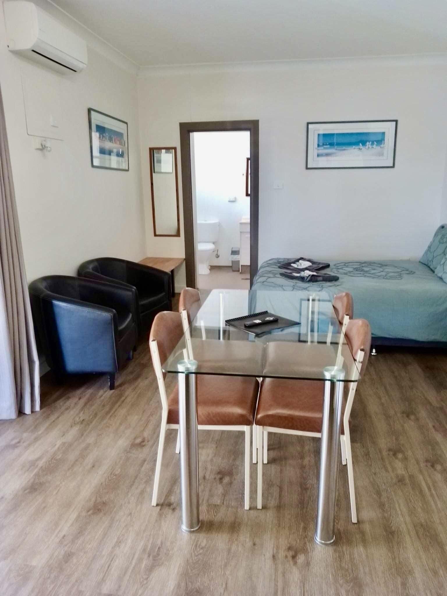 Habitación de hotel con una mesa de cristal, dos sillones de cuero, una cama con cubrecama azul, un escritorio pequeño y un ventanal con vistas.