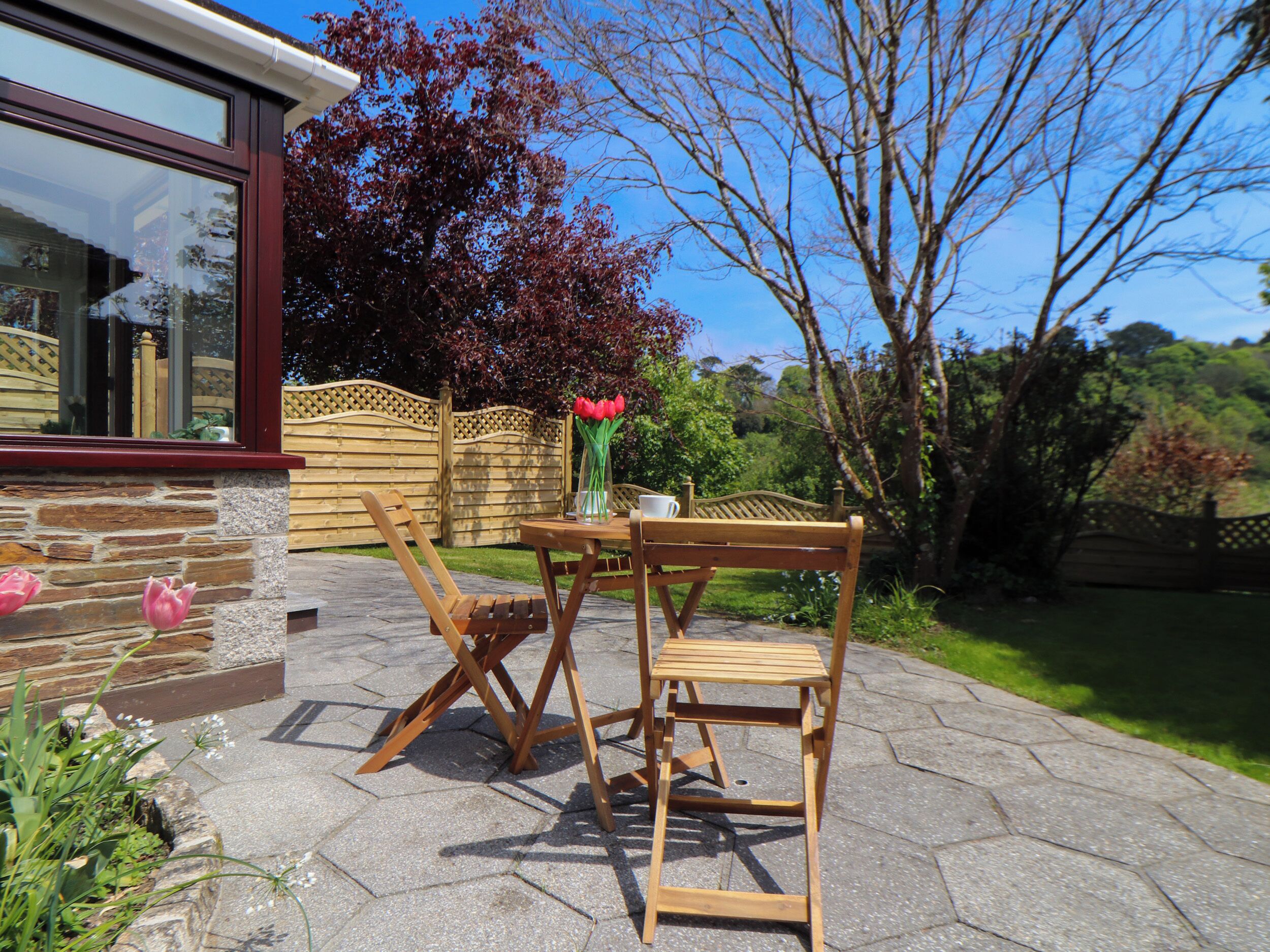 Une terrasse extérieure avec une table et des chaises en bois, un vase rempli de fleurs et un mur en pierre.