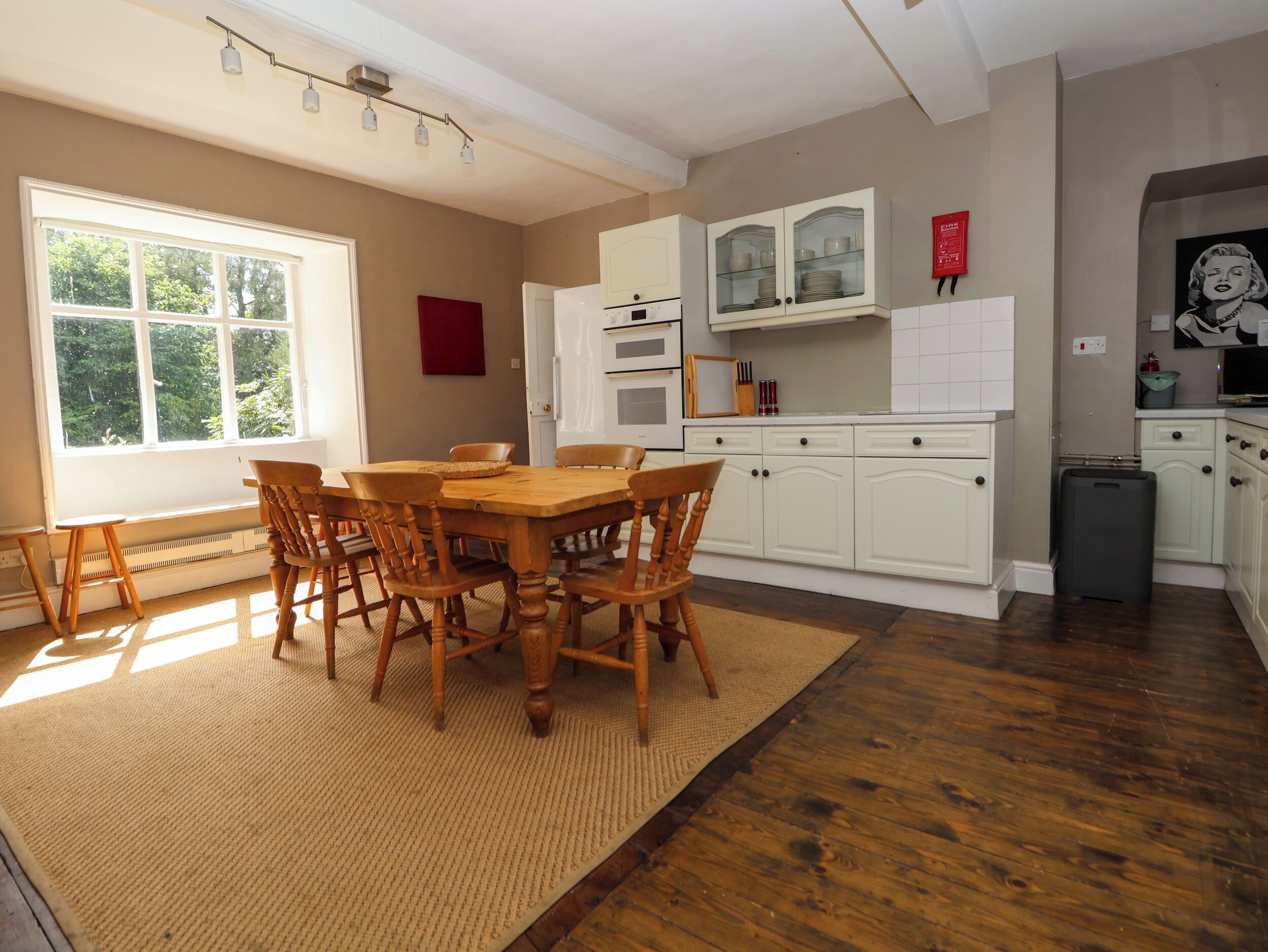 A dining area with a wooden table and chairs, a kitchen with white cabinets, and a window with a view of greenery.