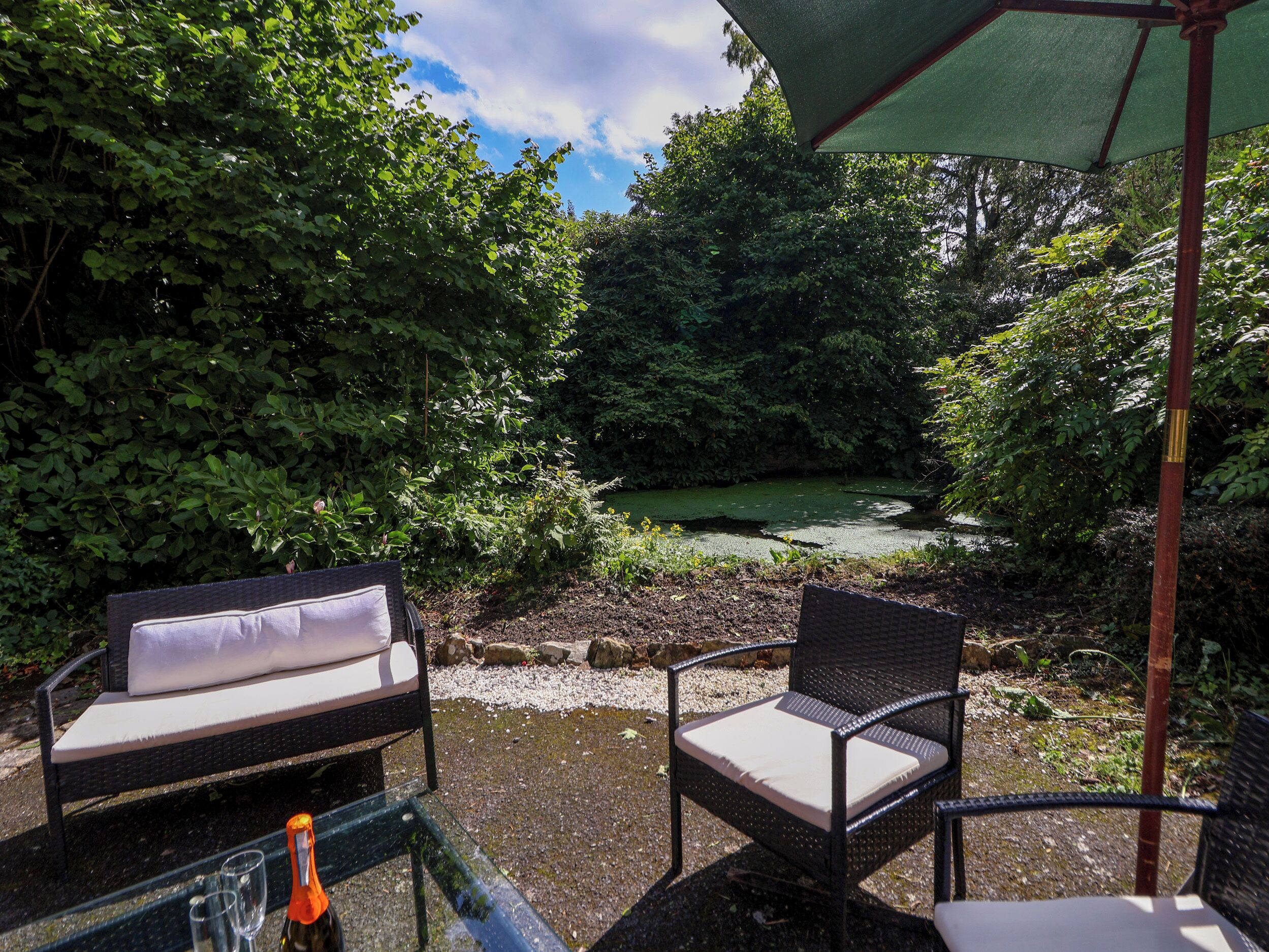 A patio with two wicker chairs and a table, overlooking a pond surrounded by dense foliage.