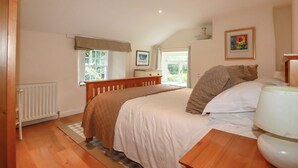A bedroom with a wooden bed, a bedside table, a radiator, and a window with a view of greenery. - Willow View (Wadebridge)