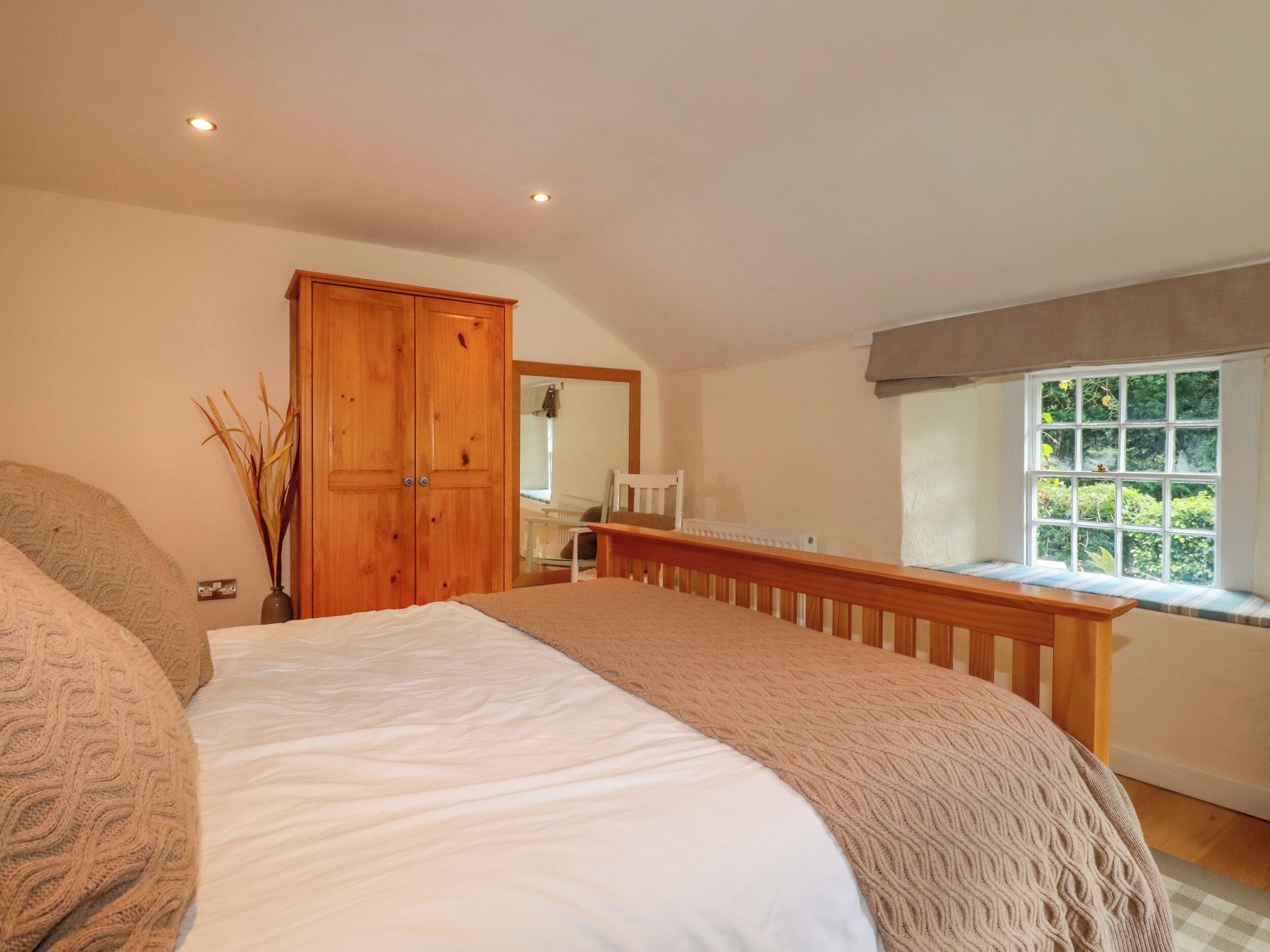 A bedroom with a large bed, wooden wardrobe, a window with a view of greenery, and a small table with a chair.