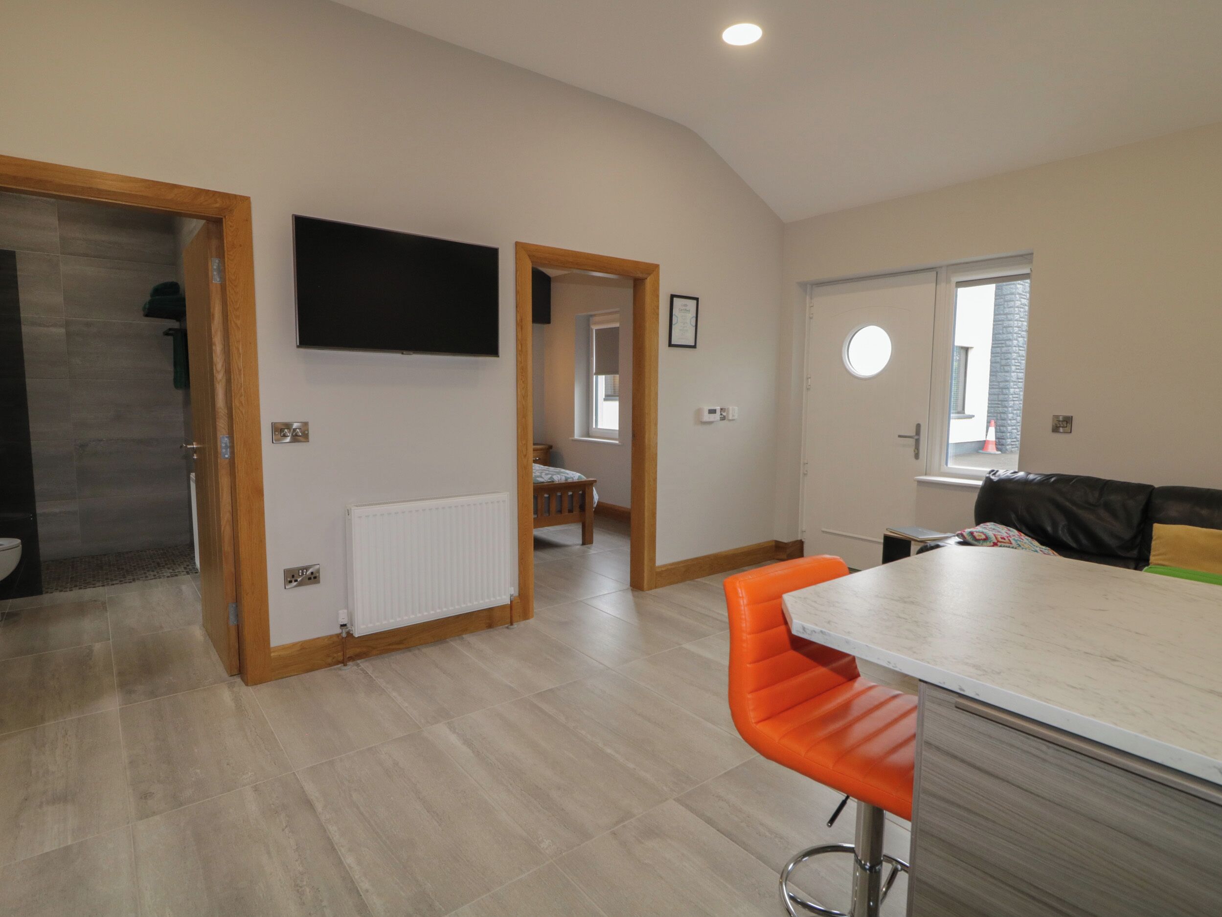 A modern kitchen with a built-in oven, a dining area with a table and orange chairs, and a living area with a sofa and a TV mounted on the wall.