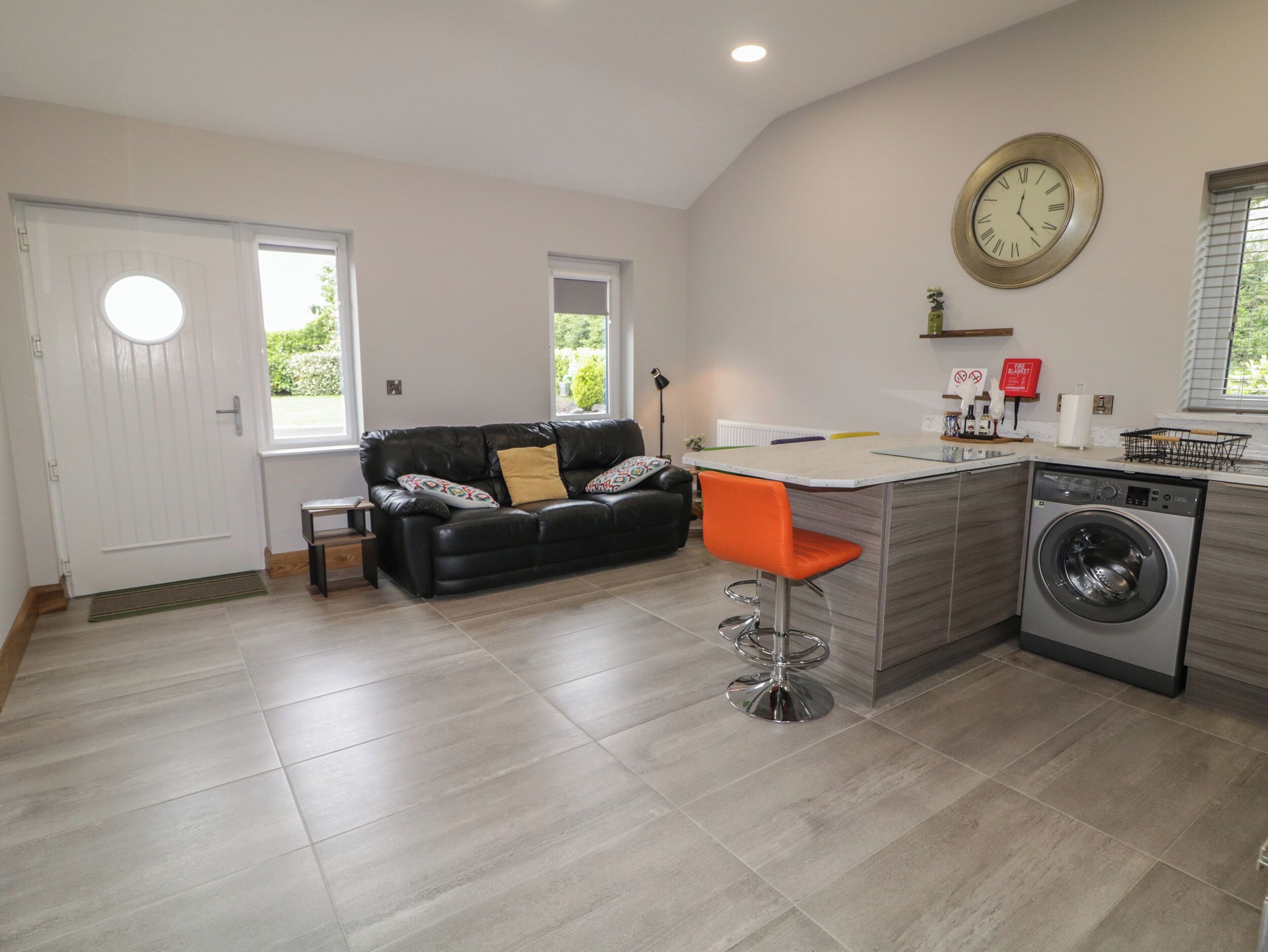 A modern kitchen with a built-in washing machine, a dining area with a black leather sofa, and a small bar stool.