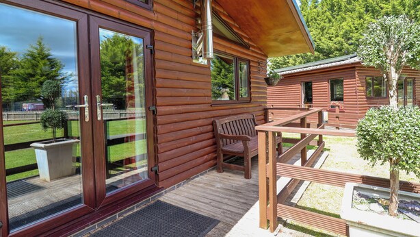 A wooden cabin with large sliding glass doors, a wooden deck, and a bench. - The Roe (Saint Asaph)