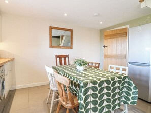 A dining area with a table covered in a patterned tablecloth, wooden chairs, a potted plant, and a refrigerator.