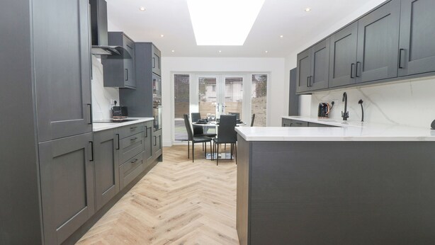A modern kitchen with dark cabinetry, a central island, and a dining area with a table and chairs. - Park View (Llandudno)