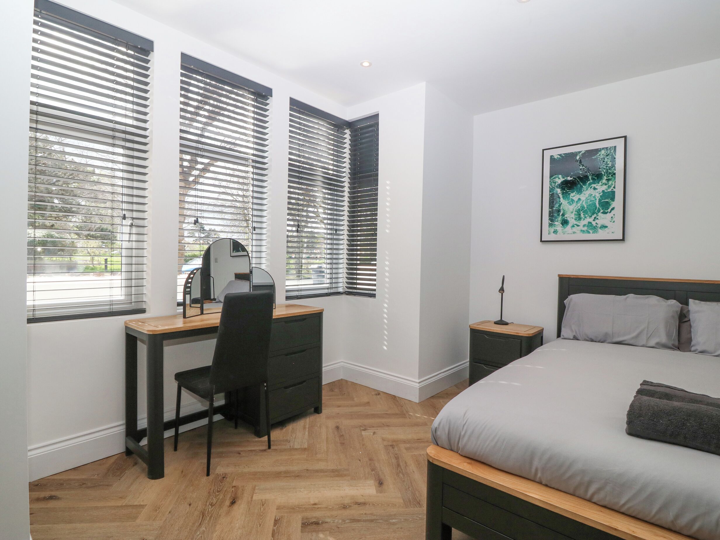 A bedroom with a bed, a desk with a chair, a bedside table, and a window with blinds.