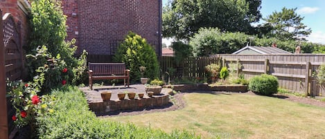 Un bâtiment en briques, avec une clôture en bois, un banc et un jardin où poussent différentes plantes et fleurs.