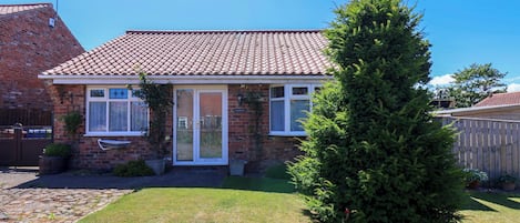 A two-story brick house with a tiled roof, a front door, and a small front yard with a lawn and a tall coniferous tree.