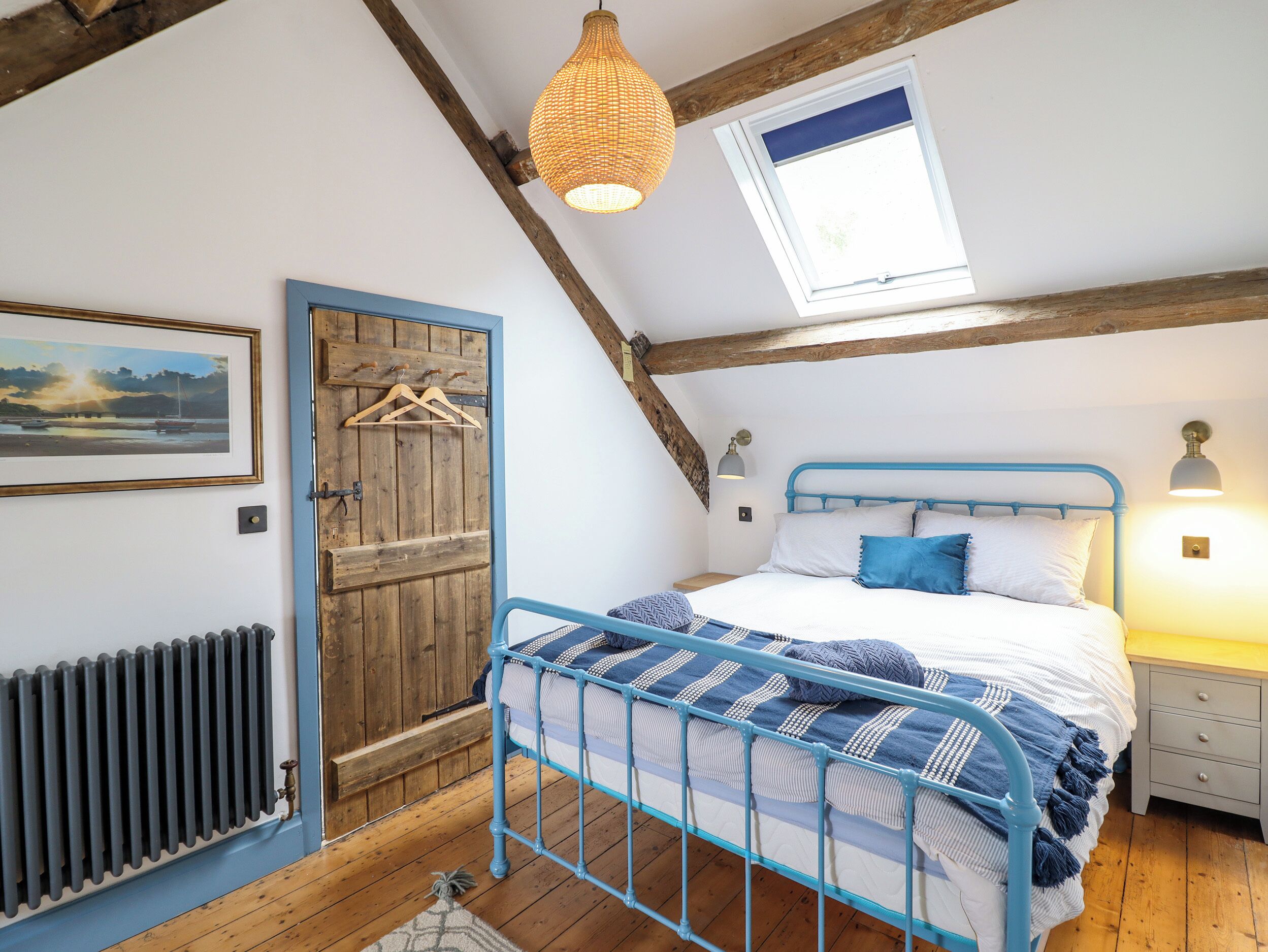 A bedroom with a blue metal bed frame, a wooden door, a radiator, a hanging light fixture, and a window with a view of the sky.