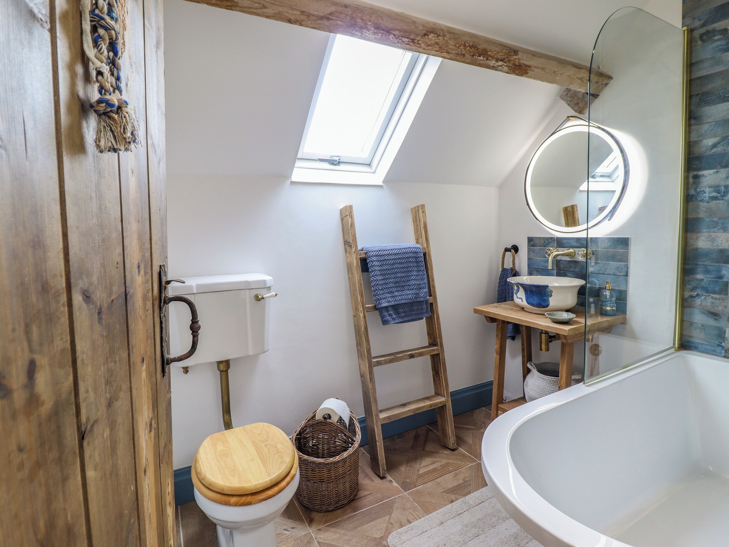 A bathroom with a skylight, wooden ladder, a toilet with a wooden seat, a bathtub, and a sink with a blue basin.