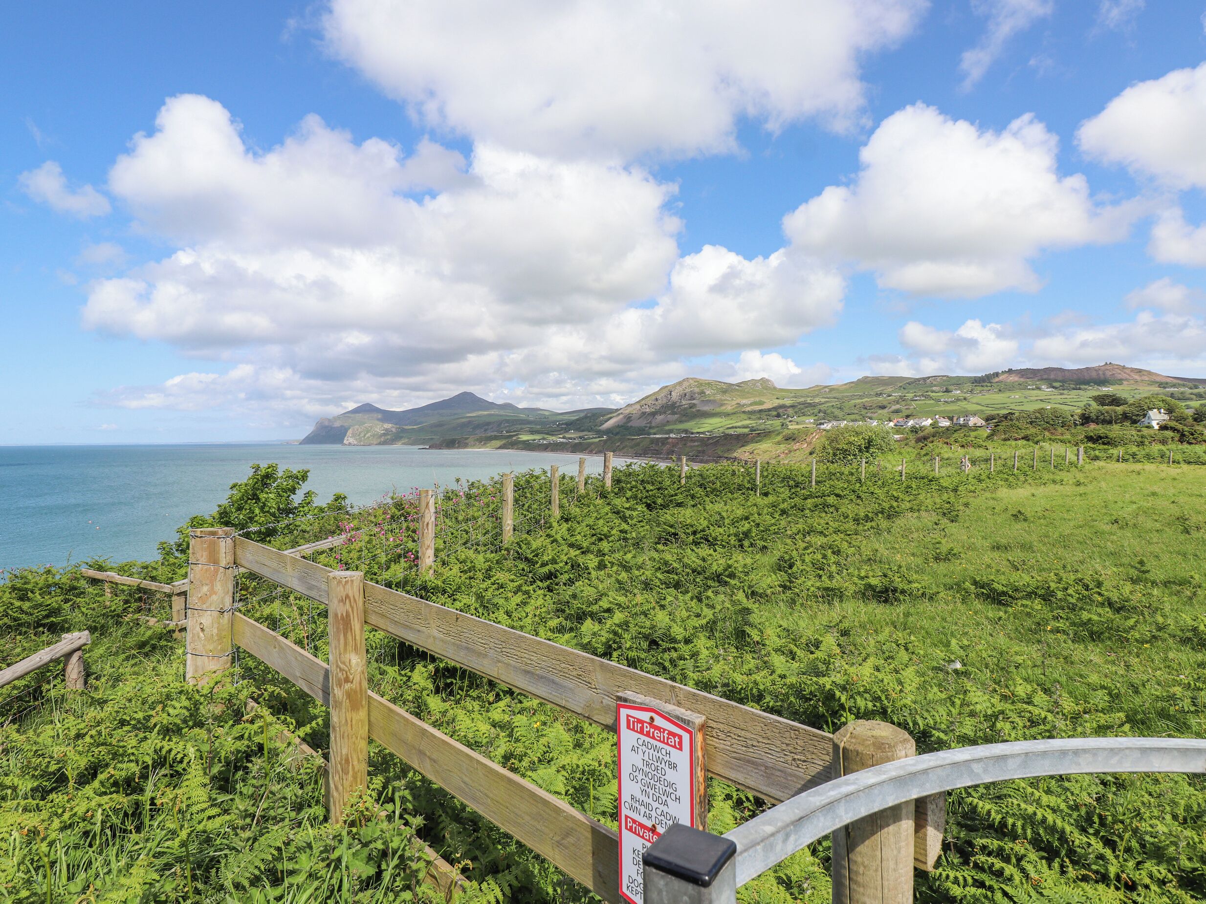 A coastal landscape with a wooden fence, a sign indicating private property, and a view of the sea and hills in the background.