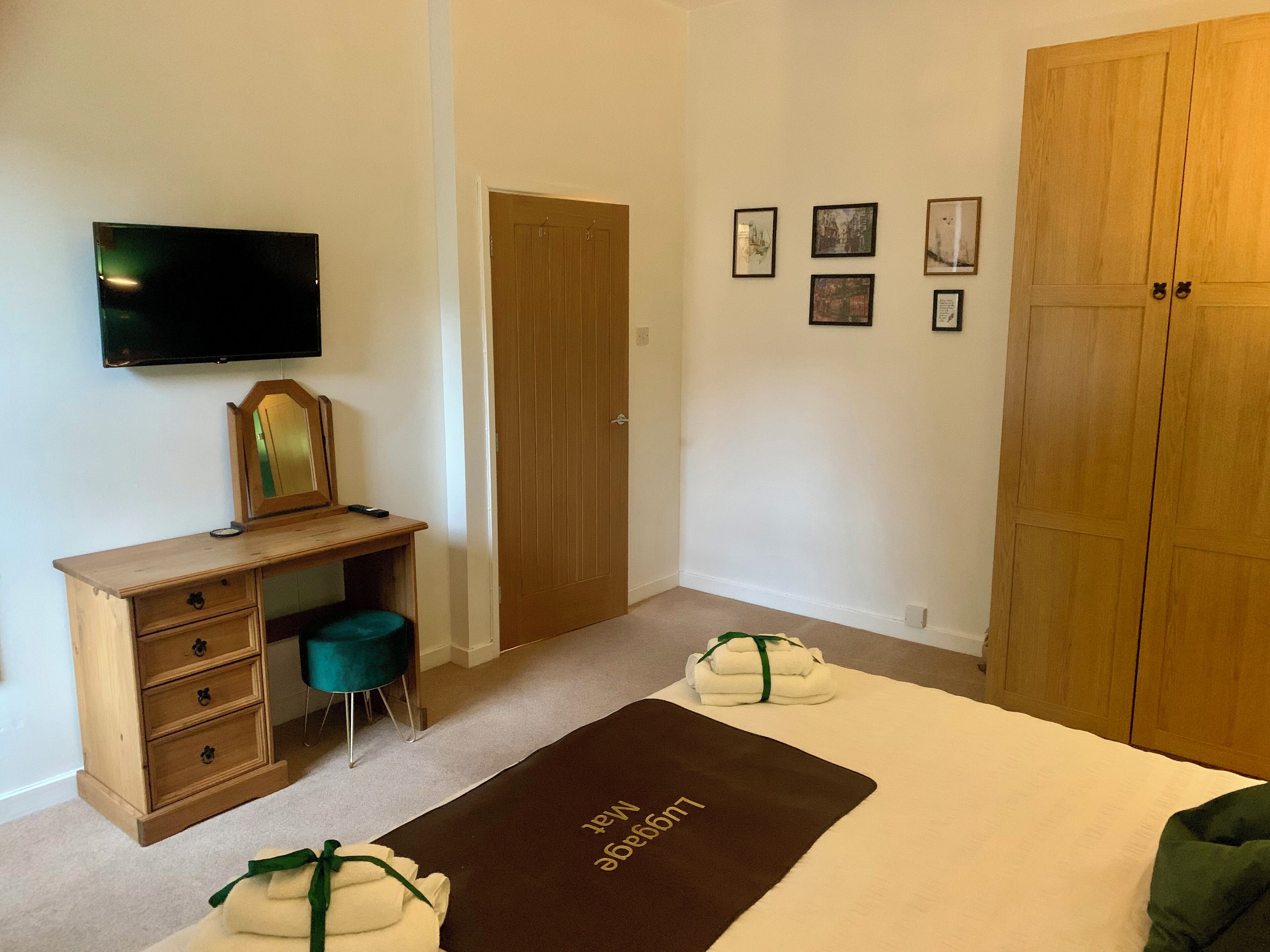 A bedroom with a wooden desk, a television, a wardrobe, a bed with a brown mat, and four framed pictures on the wall.