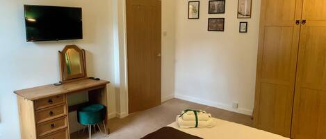 A bedroom with a wooden desk, a television, a wardrobe, a bed with a brown mat, and four framed pictures on the wall.