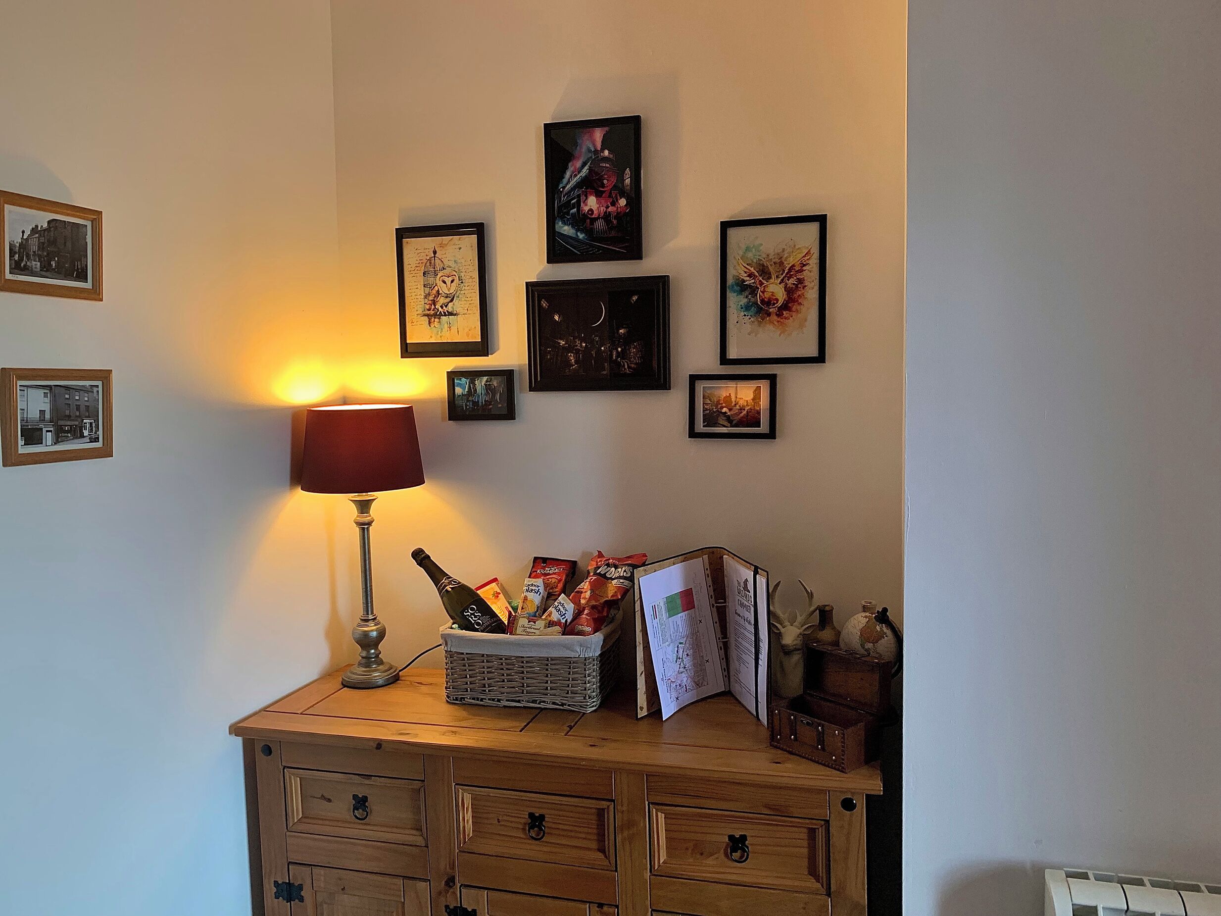 Une commode en bois avec des tiroirs, une lampe, un panier rempli de collations et des photos encadrées accrochées au mur.