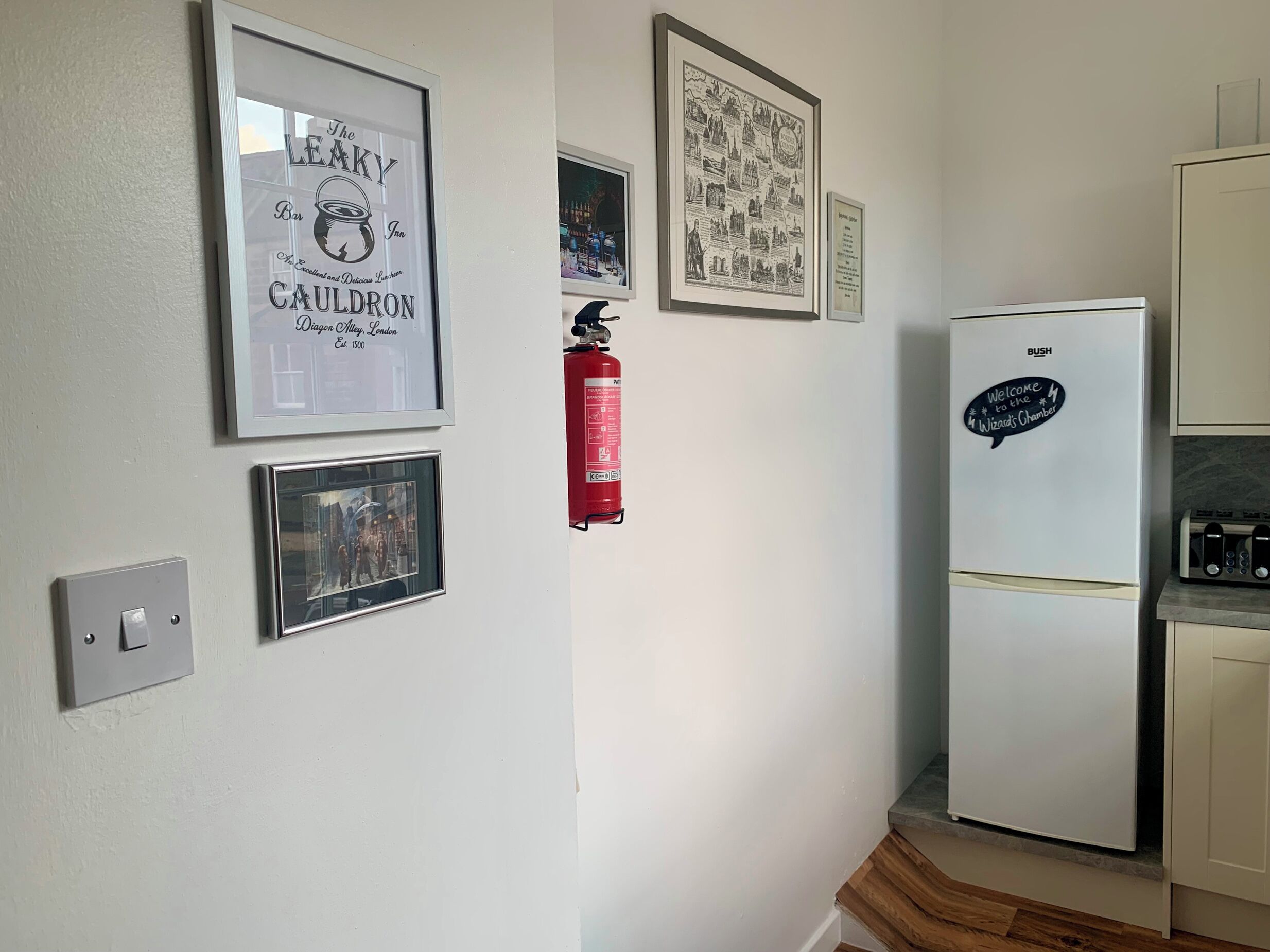 A kitchen with a refrigerator, a fire extinguisher, and framed pictures on the wall.