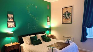 A neatly made bed with white linens and green pillows, two bedside tables with lamps, a radiator, and a framed coat of arms on the wall. - The Wizard's Chamber (Alnwick)