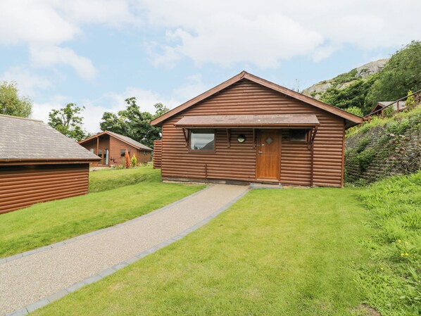 A wooden cabin with a gravel path leading to the entrance, surrounded by greenery and hills. - Luxury Lodge 62 (Rhyl)