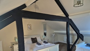 A bedroom with a sloped ceiling, a bed with white bedding, a bedside table with a lamp, a floor lamp, and a small bedside table with a mirror. - 12 Eagle Parade (Buxton)