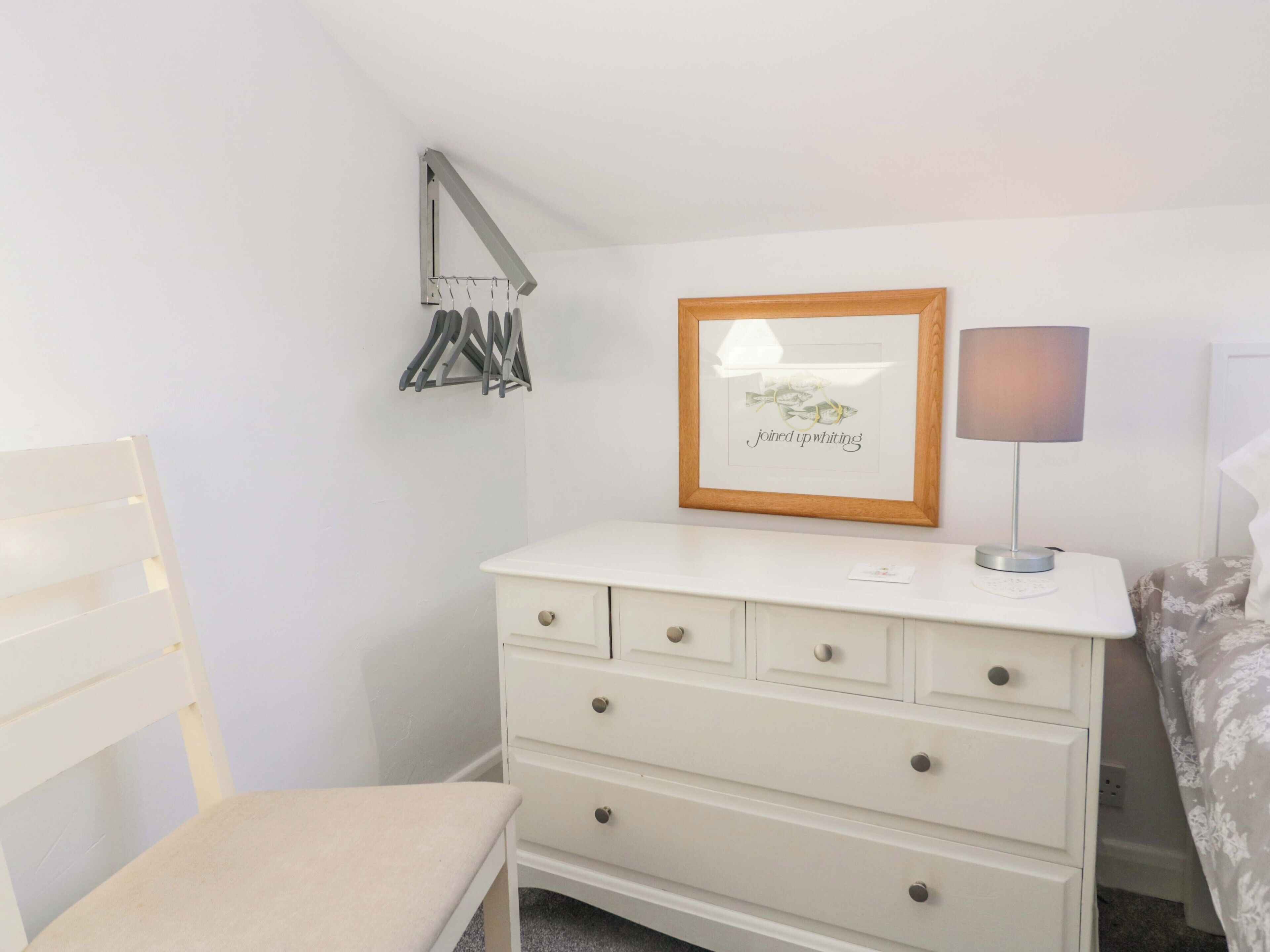 A bedroom with a white dresser, a chair, a framed picture on the wall, and a bedside lamp.