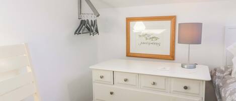 A bedroom with a white dresser, a chair, a framed picture on the wall, and a bedside lamp.