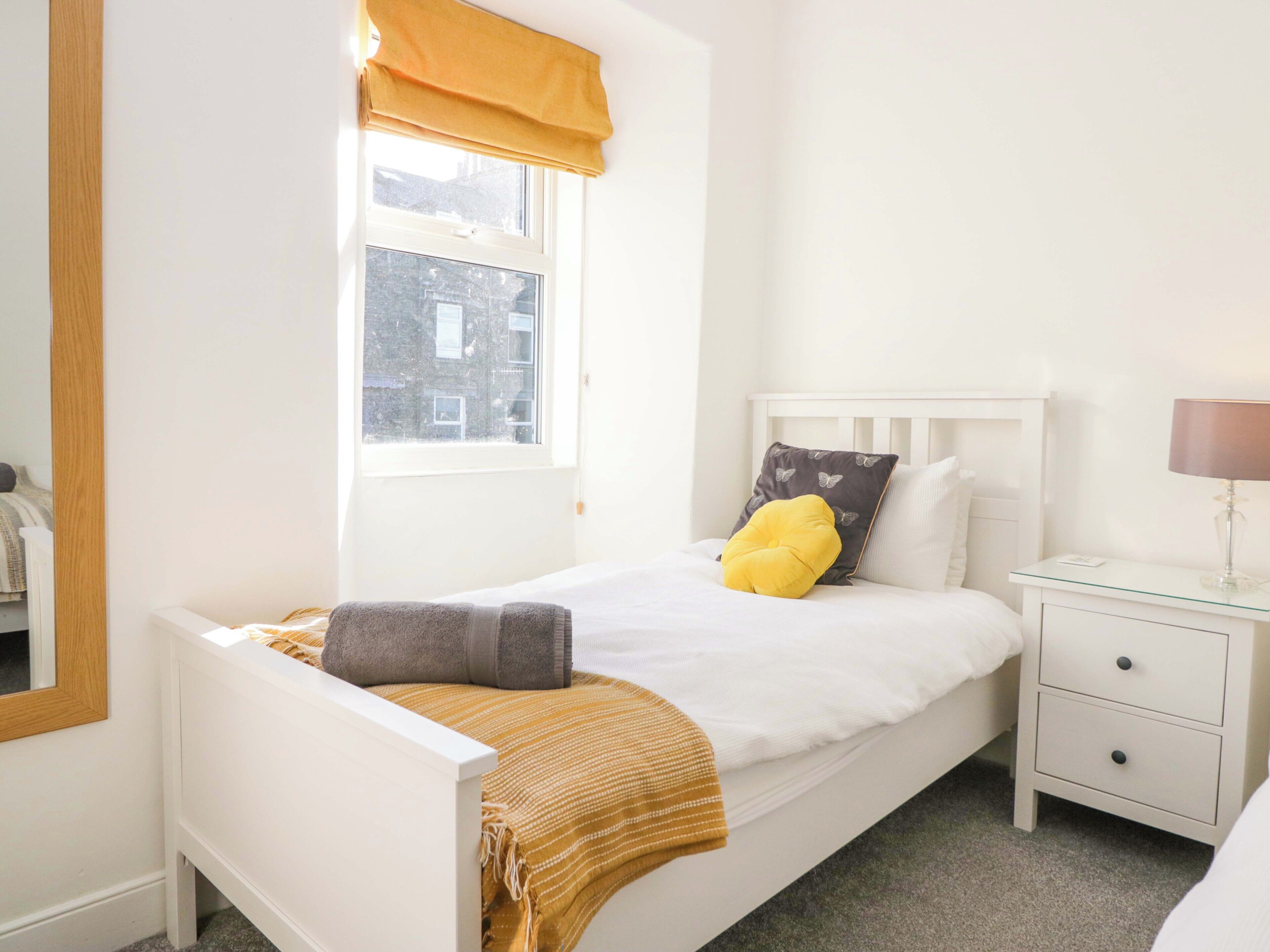Une chambre à coucher bien rangée, avec un lit simple, une table de chevet avec une lampe et une fenêtre ornée d’un rideau jaune.