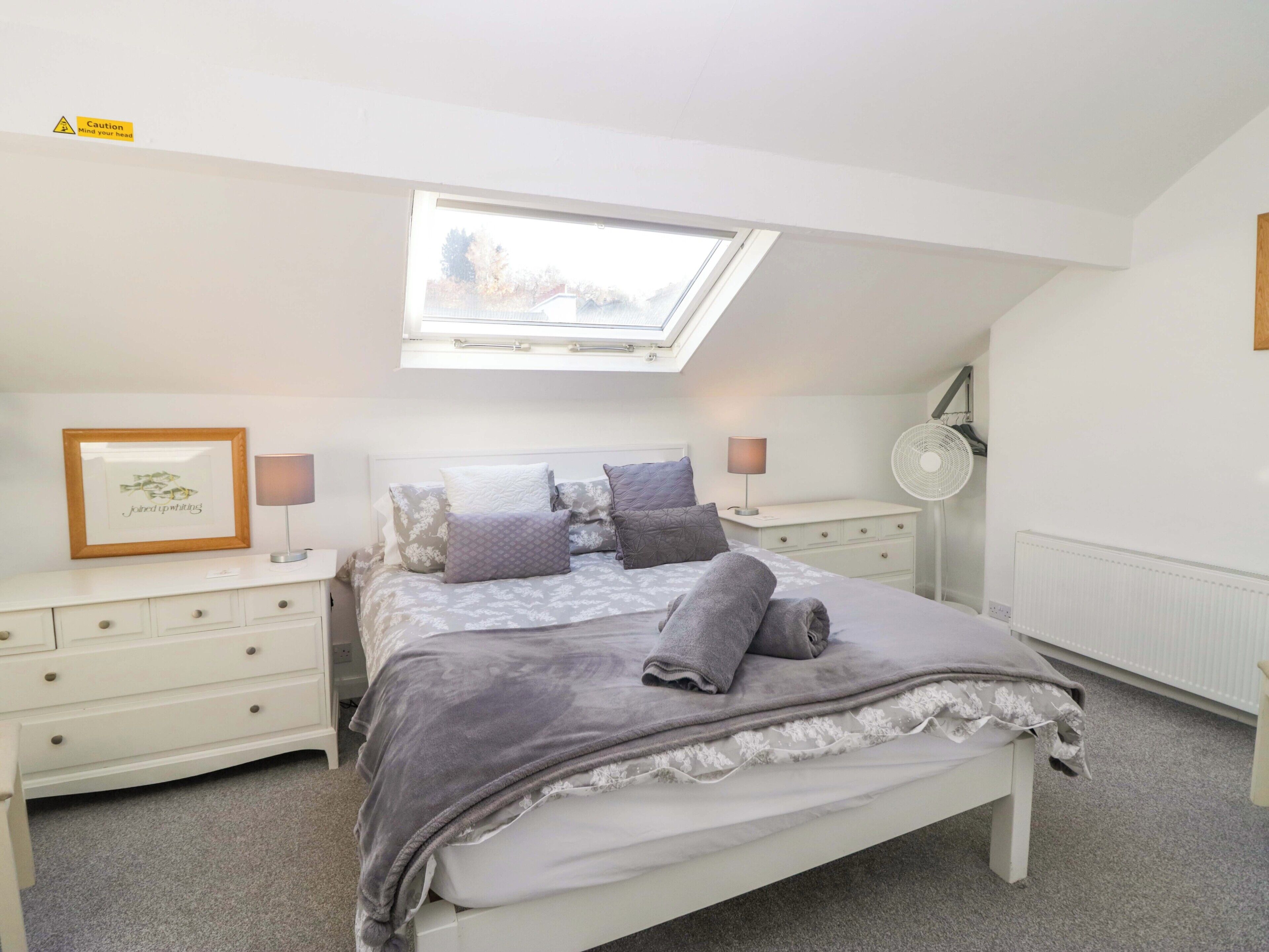 A bedroom with a skylight, a bed with a grey blanket, a bedside table with a lamp, and a framed picture on the wall.