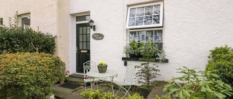 A white cottage with a black door, a small garden with a table and chairs, and a sign that reads "Acorn Cottage".
