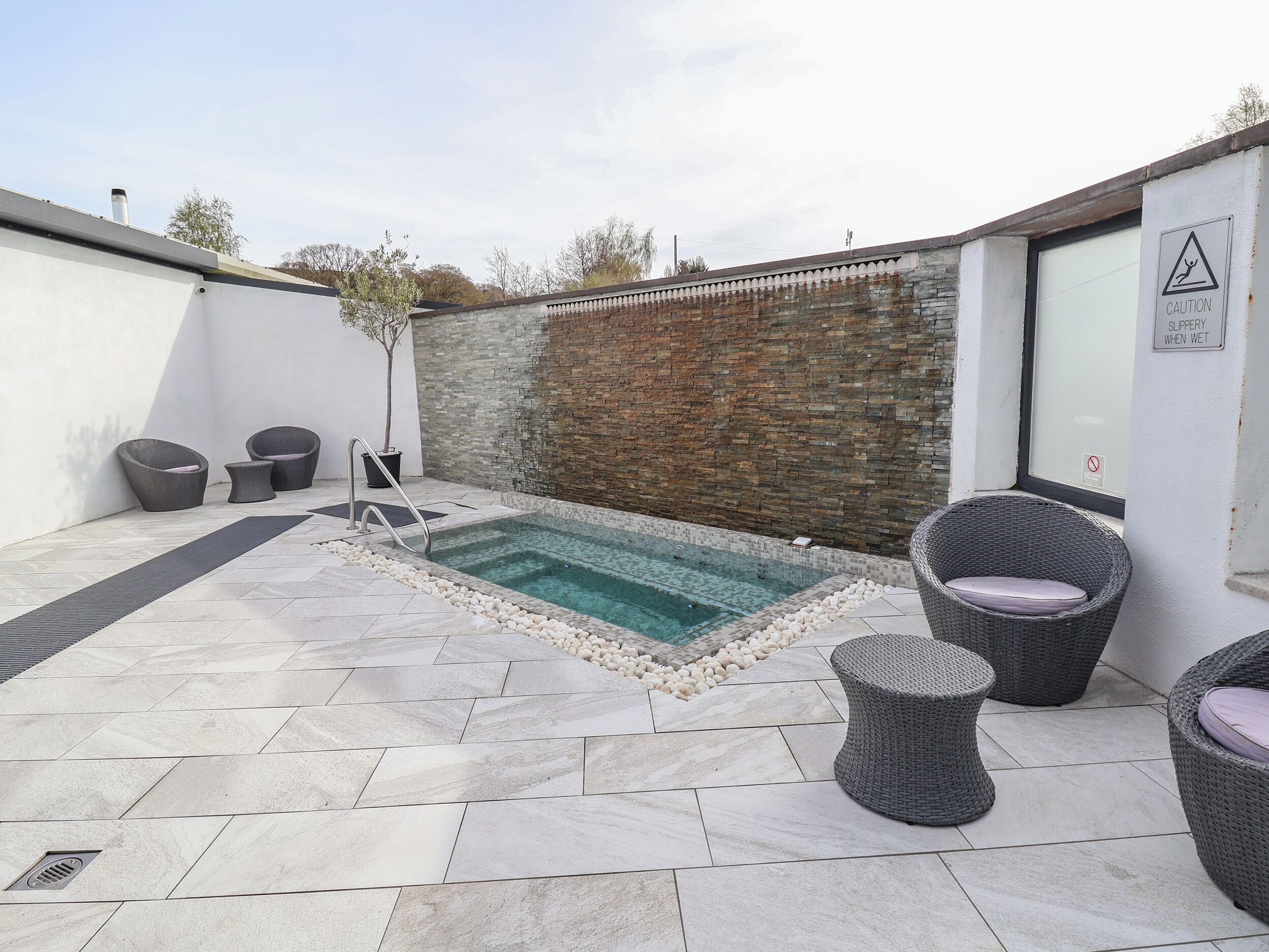 A small outdoor swimming pool with a stone border, surrounded by wicker furniture and a brick wall.