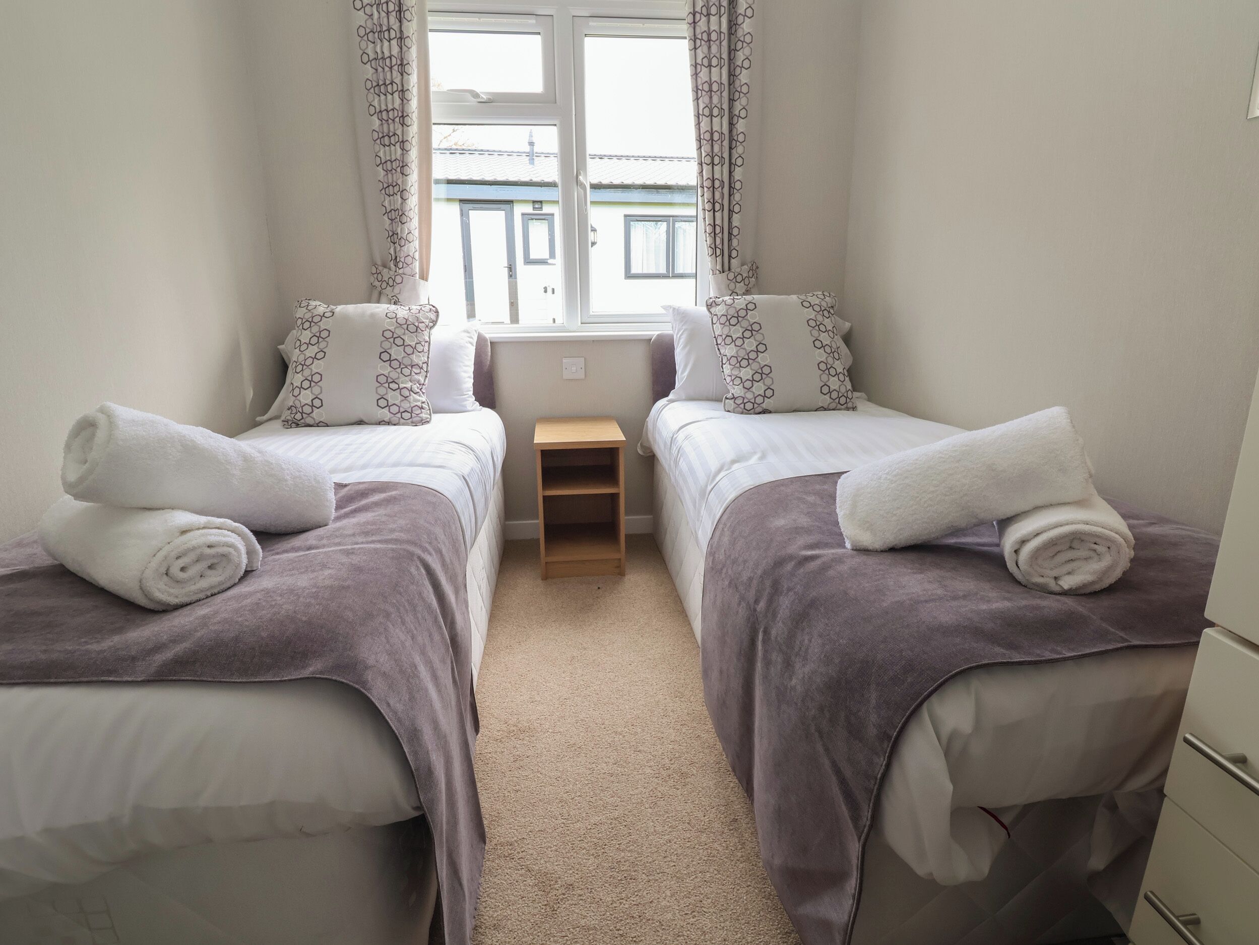 A twin room with two beds, each with a rolled towel, a small wooden nightstand, and patterned pillows.