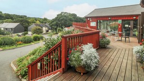 Una terraza de madera con mobiliario exterior, rodeada de exuberante vegetación y una cerca roja.
