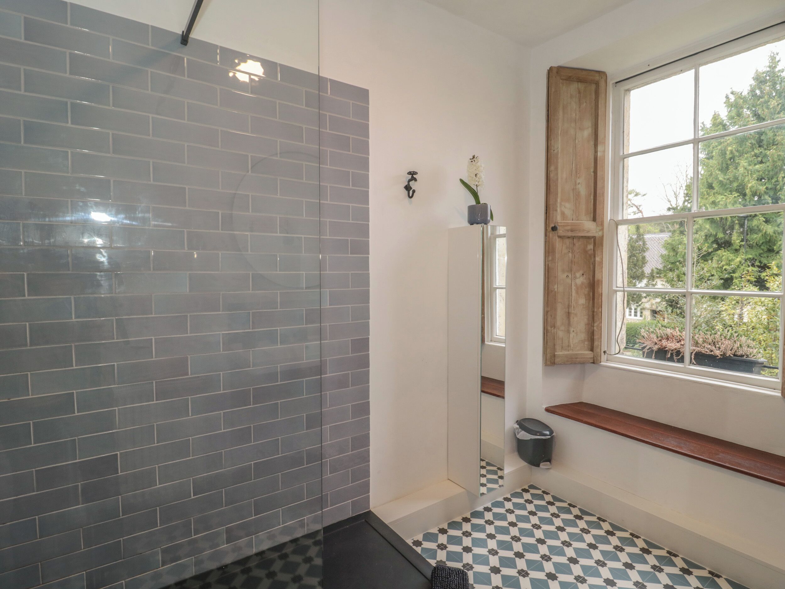 Ein Badezimmer mit grauen Fliesenwänden, einem Schachbrettfußboden, einer Holzbank, einem Fenster mit Blick ins Grüne und einem kleinen Mülleimer.