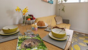 Une table à manger avec un magazine, des bols, une assiette de pain et un vase de fleurs.
