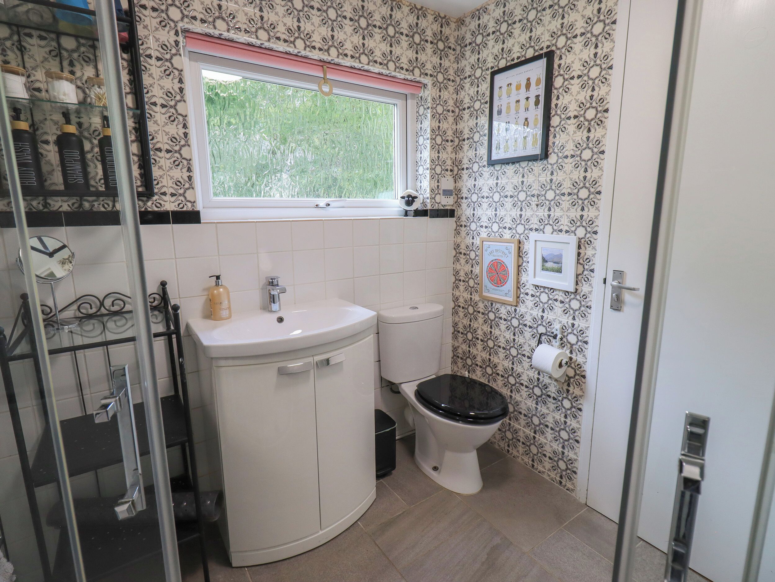 Une salle de bain avec un lavabo, des toilettes et une douche, le tout de couleur blanche. Il y a une fenêtre au-dessus du lavabo et un tableau encadré au mur.