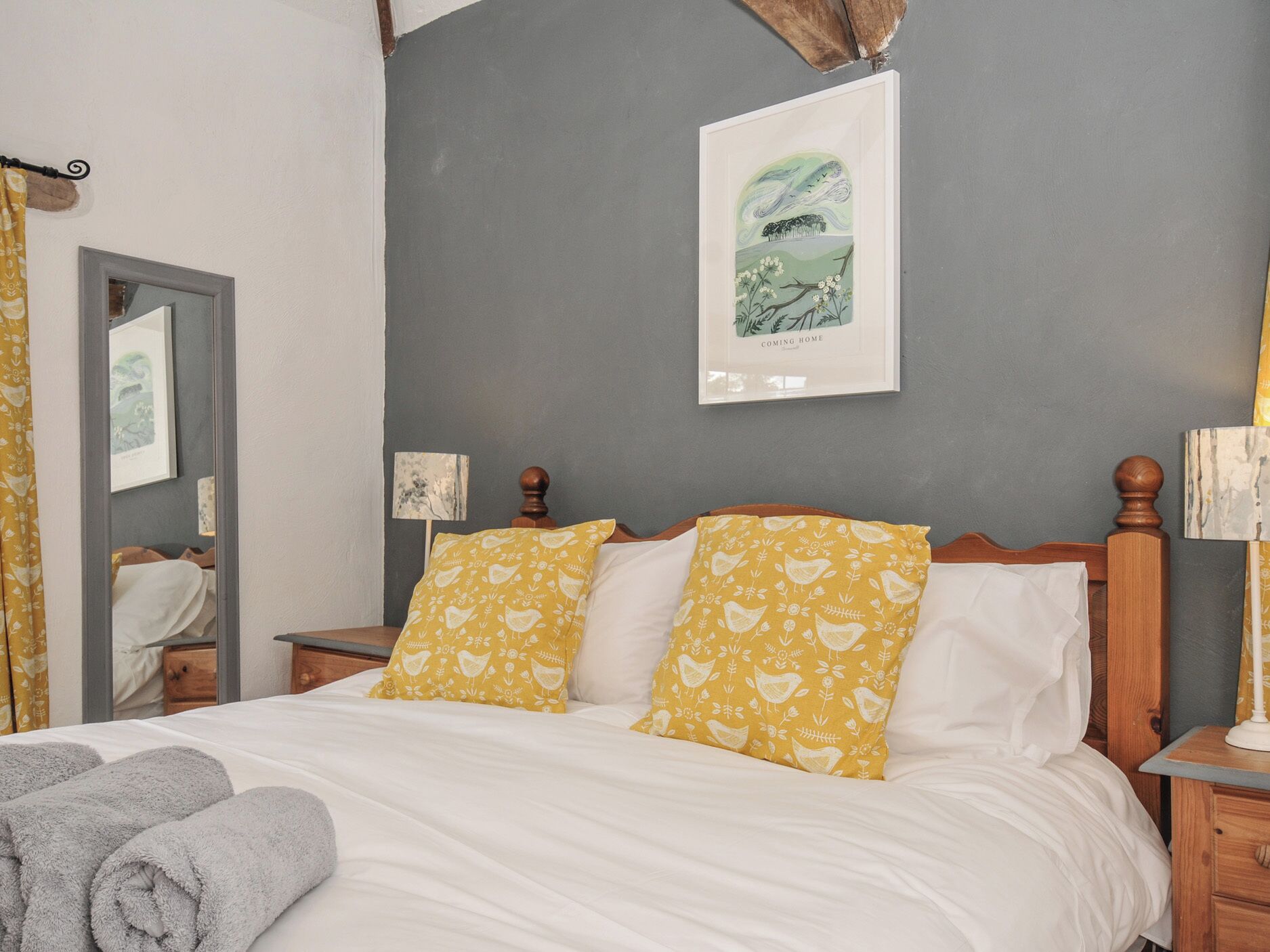 A bedroom with a bed, two pillows, a bedside table, a lamp, a mirror, and a framed picture on the wall.