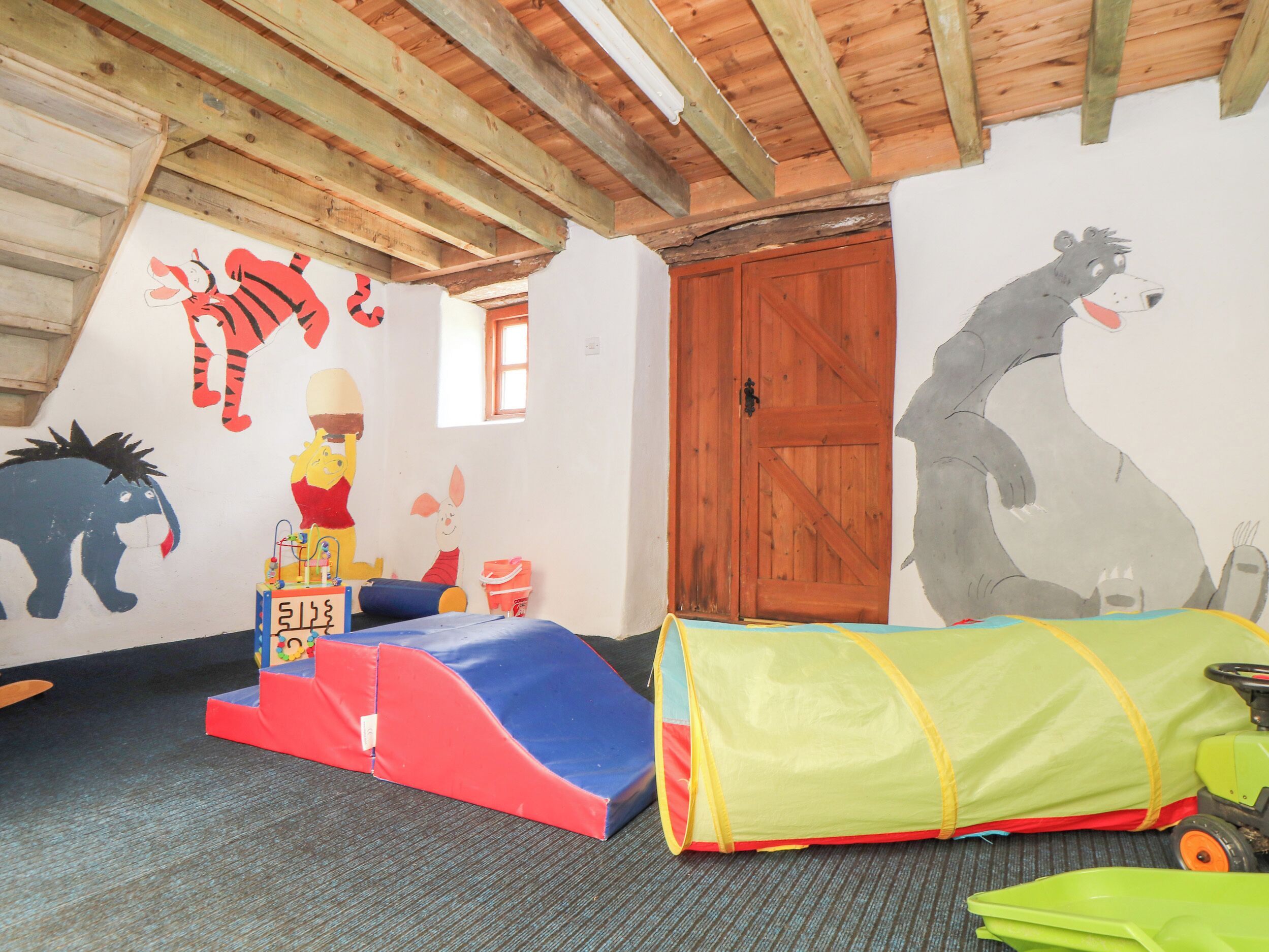 A children's playroom with a wooden ceiling, a red and blue play structure, a green tent, and wall murals of Winnie the Pooh and a hippo.