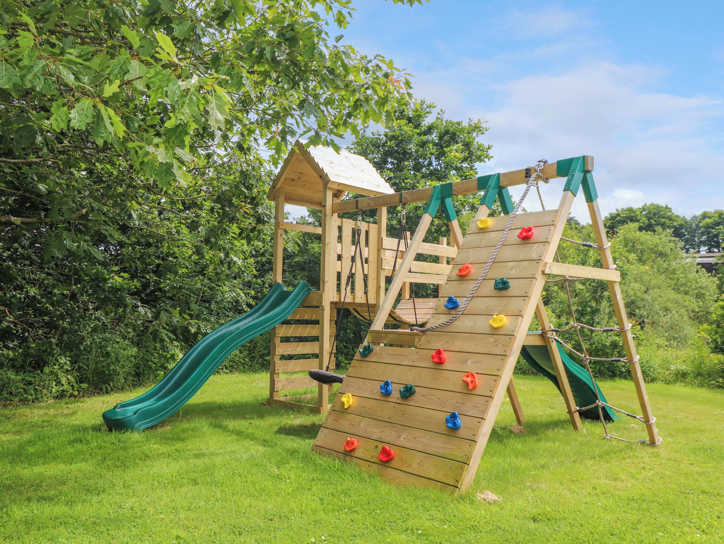 A wooden play structure with a slide and climbing wall, surrounded by grass and trees.