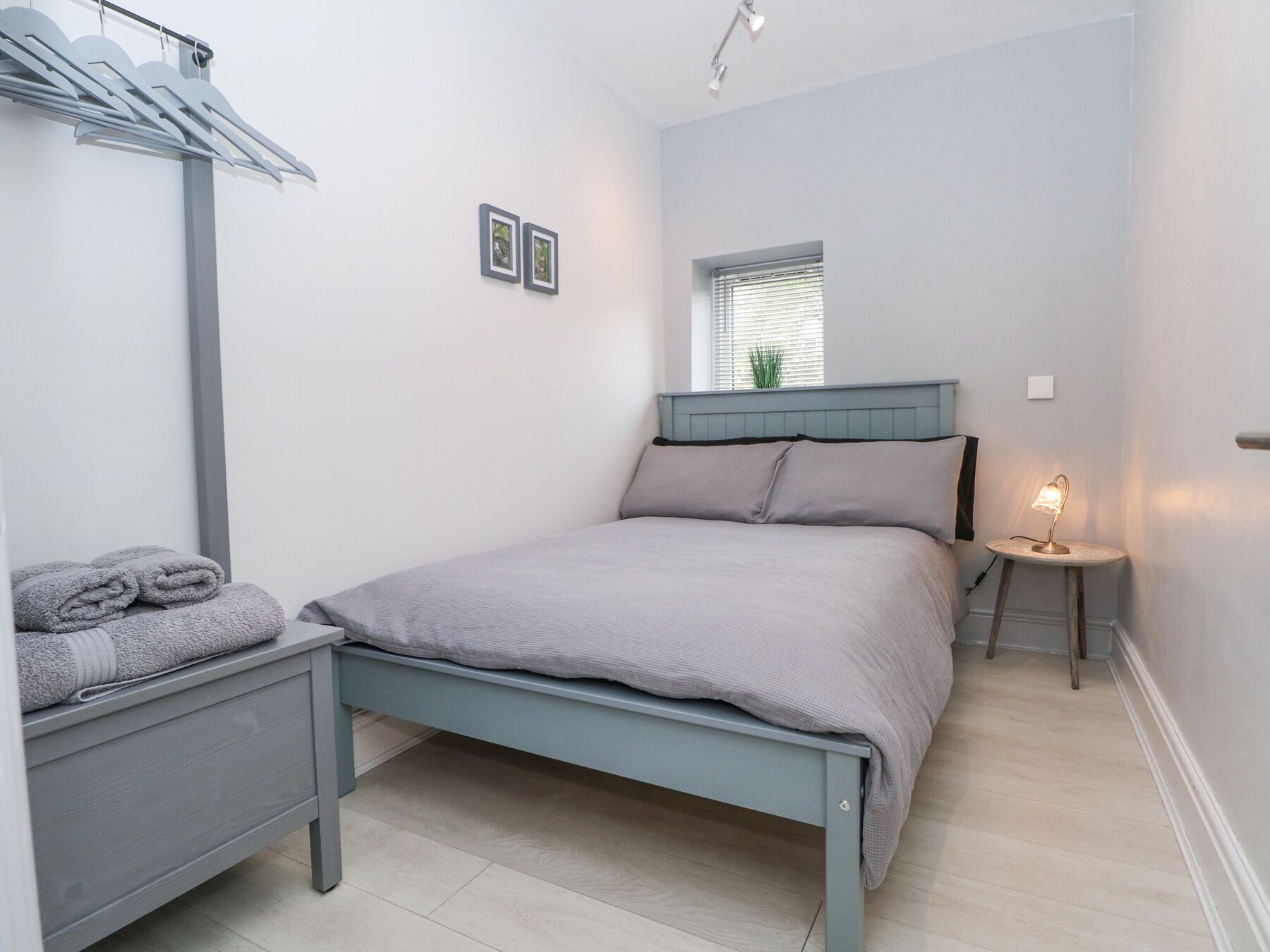 A bedroom with a bed, a bedside table, a lamp, a window, and a wall-mounted shelf with towels.