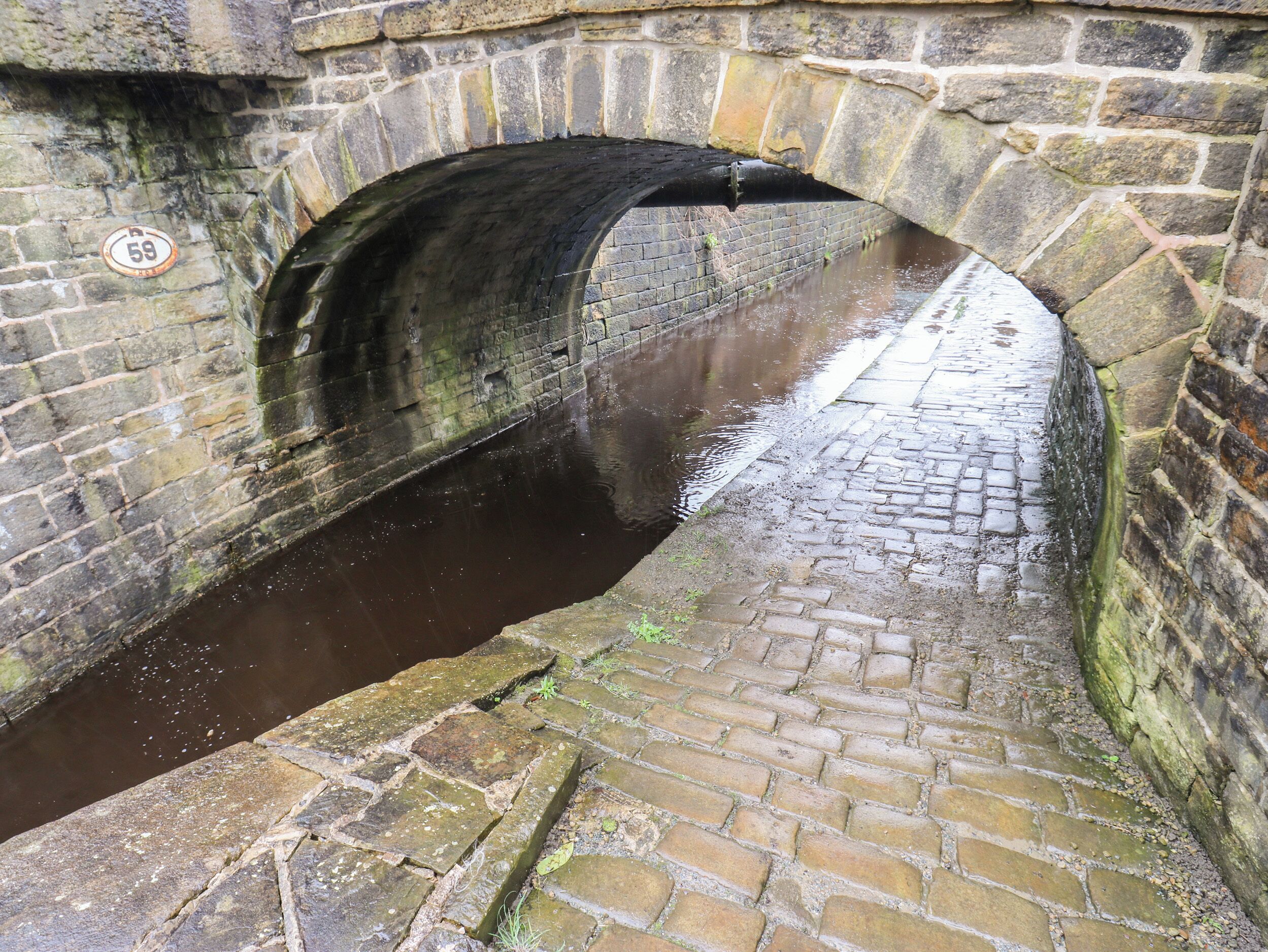 A stone bridge over a canal with a lock mechanism, a stone wall, and a sign with the number 59.