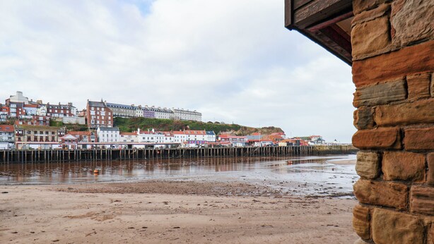 A coastal view with a stone pier extending into the water, a sandy beach, and a row of buildings with red roofs in the background. - Kitty's Cottage (Whitby)