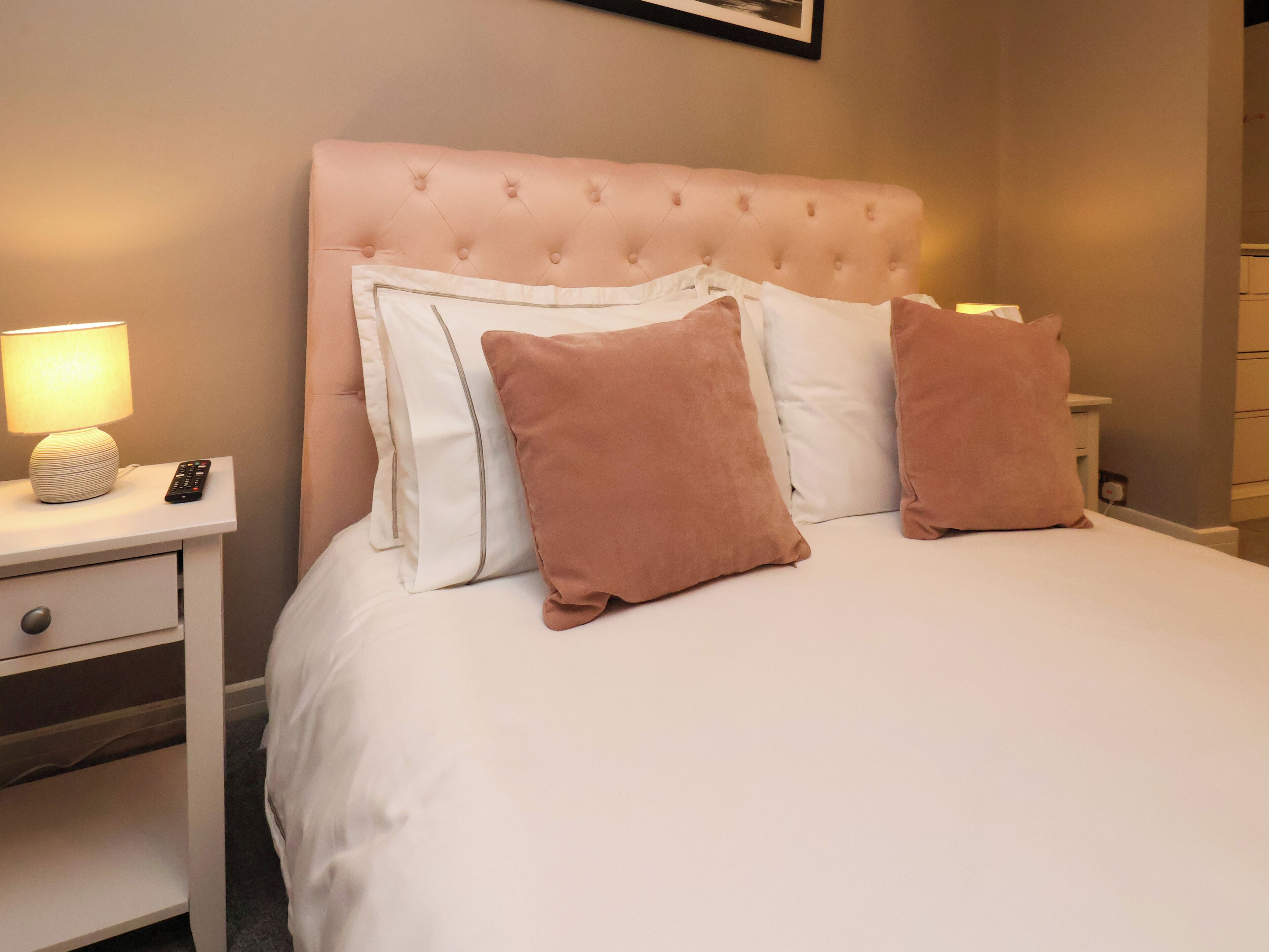 A neatly made bed with a tufted headboard, a bedside lamp, and a remote control on a nightstand.