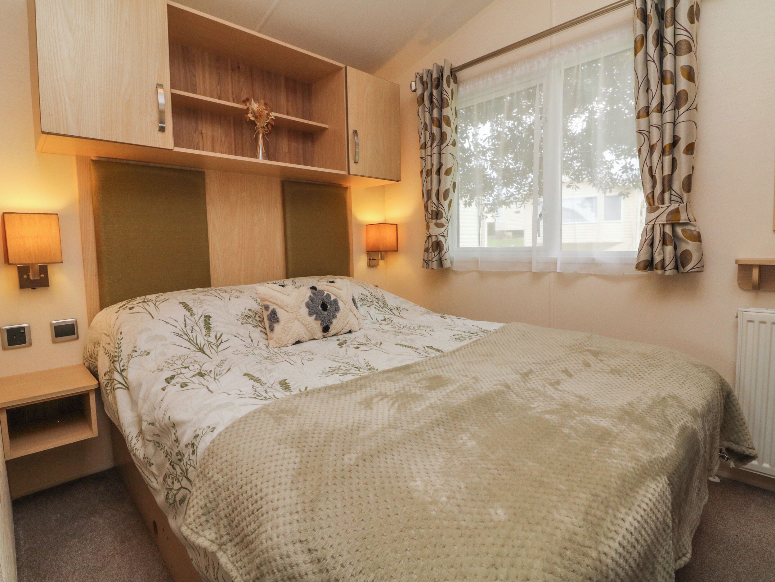 Un dormitorio con cama, cabecera de madera, mesitas de noche, una ventana con cortinas y lámparas de pared.