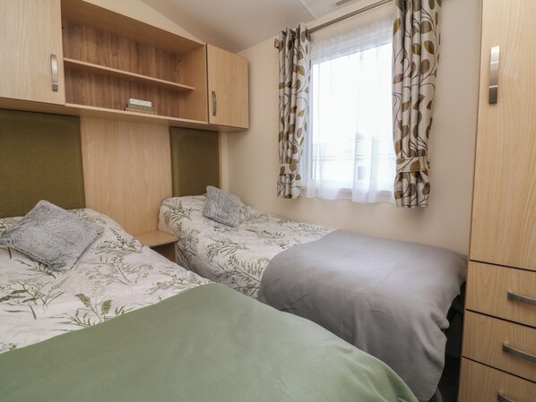 A room with two beds, wooden cabinets, and a window with patterned curtains. - Ocean View (Ilfracombe)