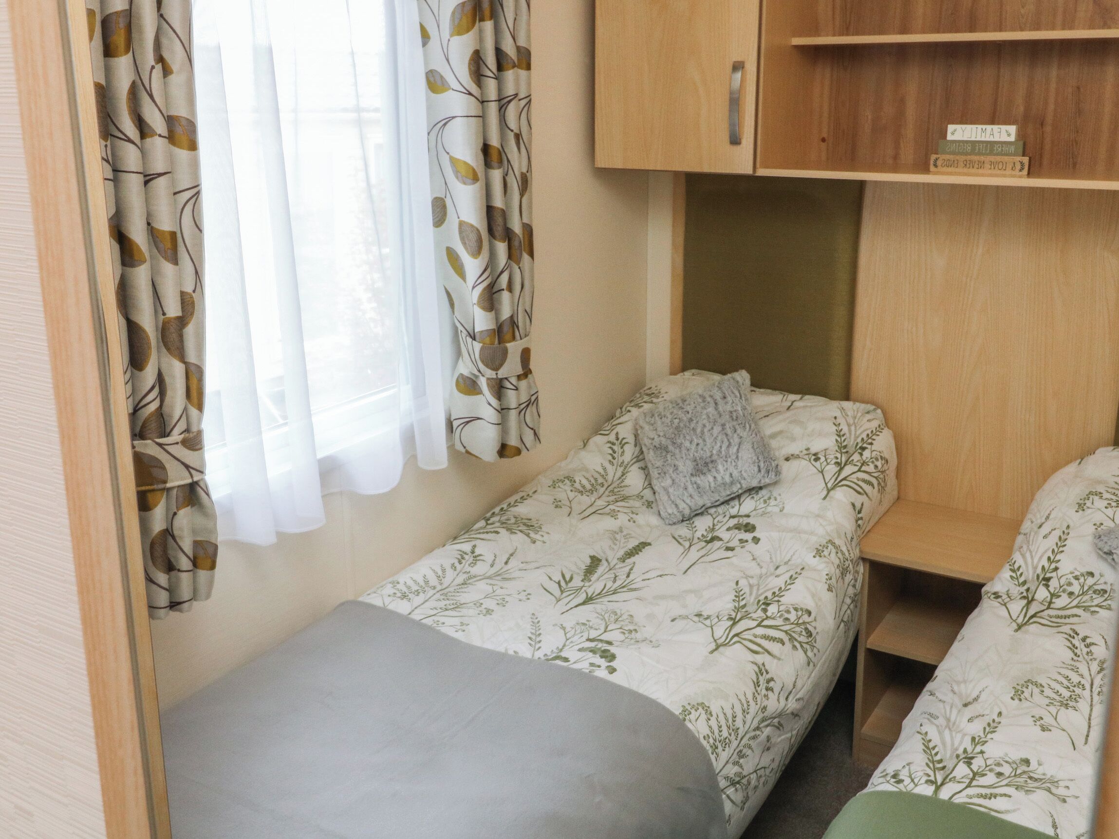 A small, compact bedroom with two single beds, a wooden wardrobe, and a window with leaf-patterned curtains.