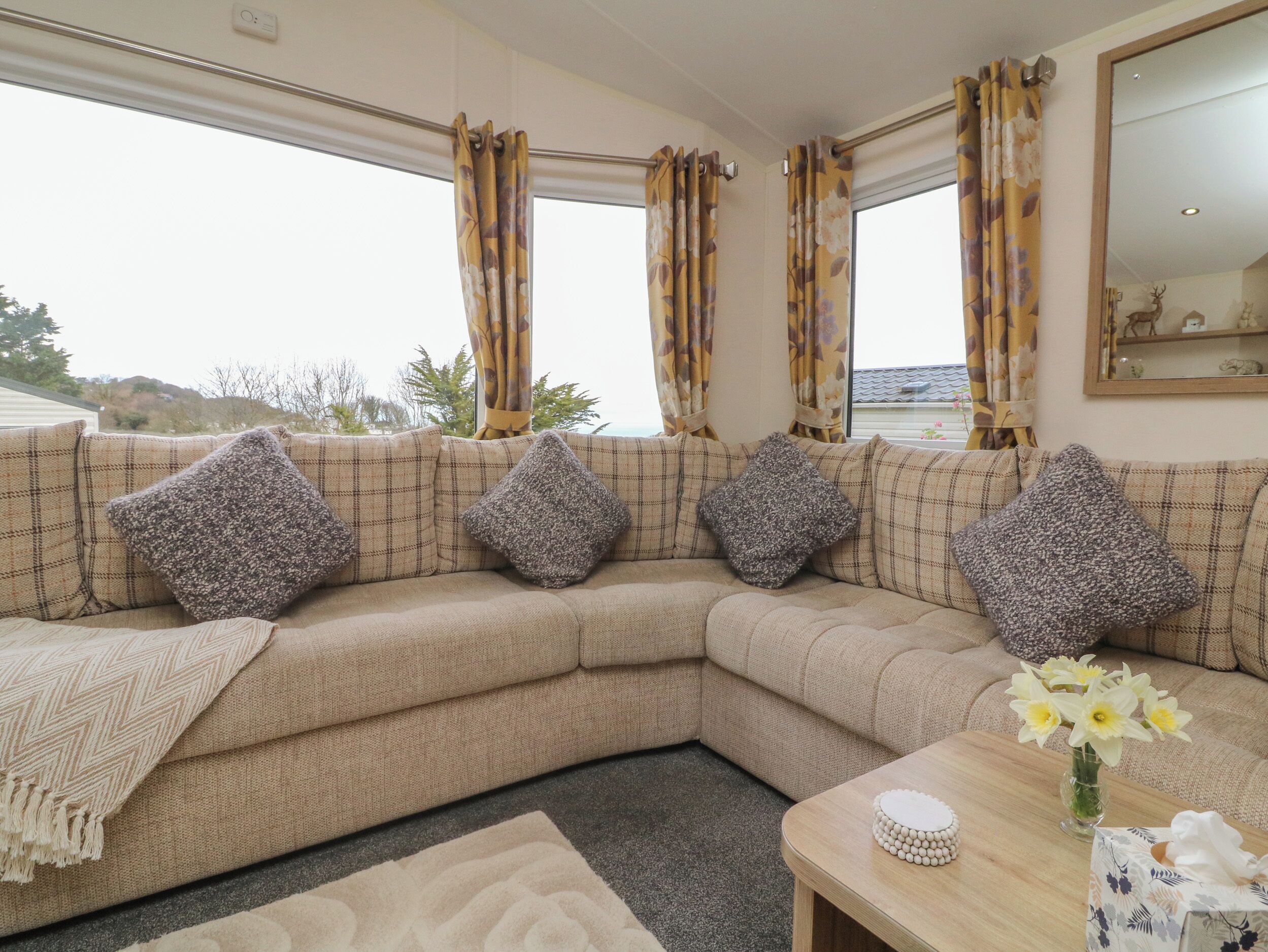 A beige sectional sofa with patterned cushions, a wooden coffee table with a vase of flowers, and a view of the outdoors through large windows with floral curtains.
