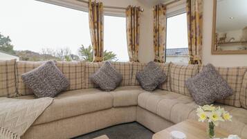 A beige sectional sofa with patterned cushions, a wooden coffee table with a vase of flowers, and a view of the outdoors through large windows with floral curtains.